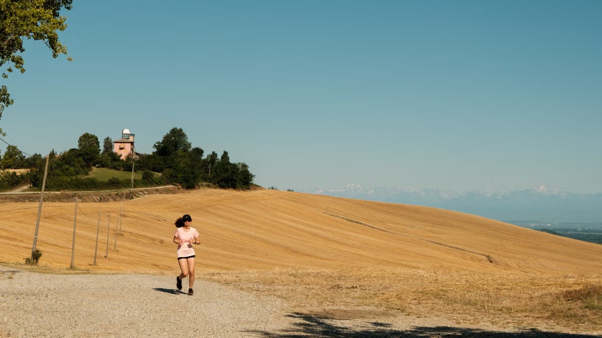 A woman runs on a gravel path through a sunlit rural landscape with dry fields and blue sky.