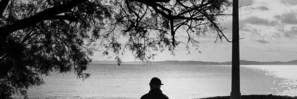 Black and white image of a person sitting on a bench by the sea, under a tree.